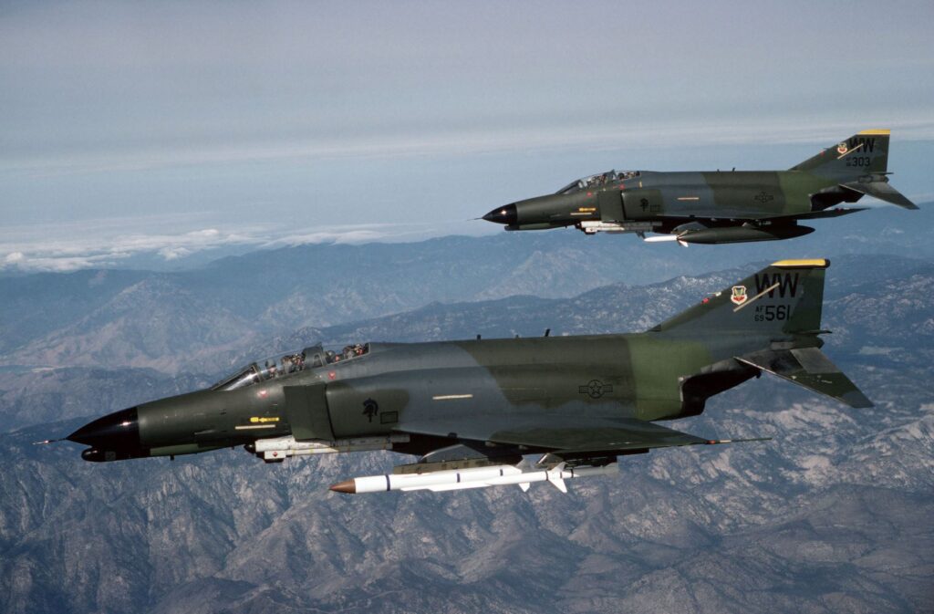 A left side view of an F-4E Phantom II and an F-4G Advanced Wild Weasel Phantom II aircraft from the 561st Tactical Fighter Squadron. Both aircraft are equipped with AN/ALQ-119 electronic countermeasures pods and AGM-45 Shrike missiles. The F-4G is also equipped with an AGM-78 Standard anti-radiation missile on the left inside wing pylon.